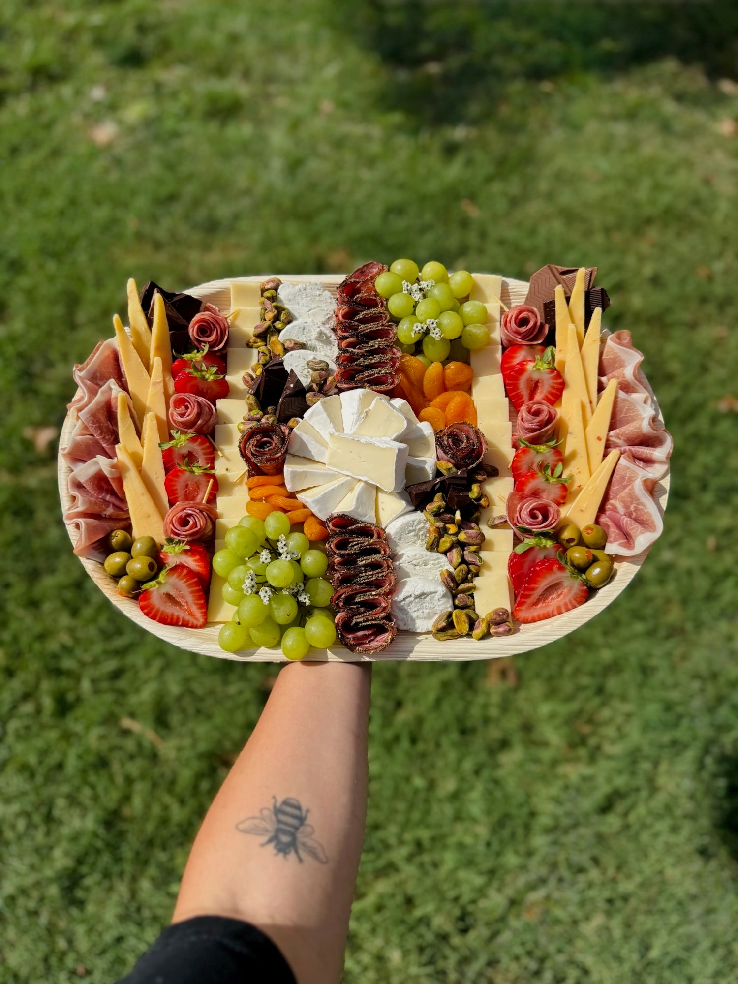 Platter of assorted meats, cheeses, and fruits held by a person outdoors.
