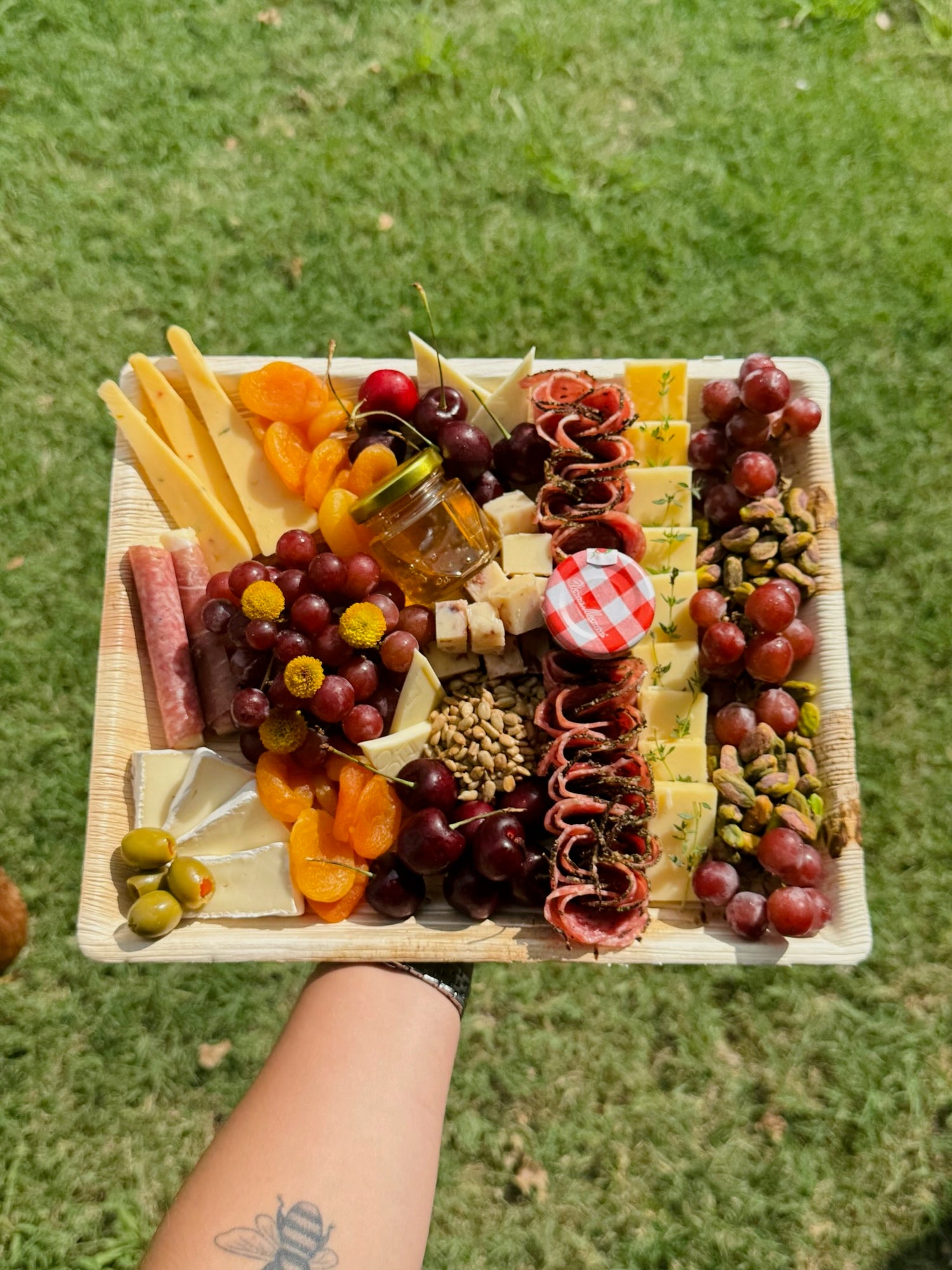 Assorted fruits, cheeses, and meats on a wooden board held over grass.
