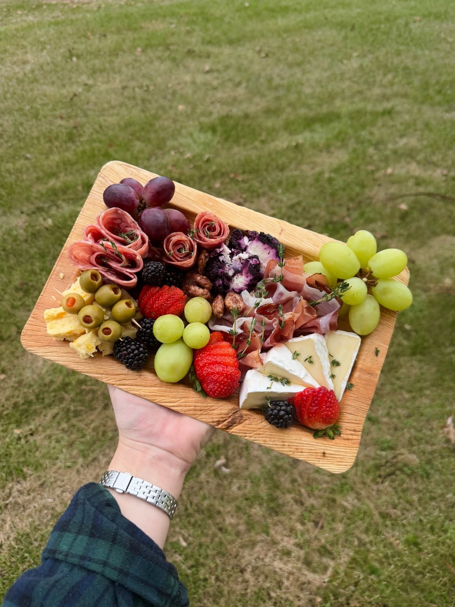 Wooden platter with assorted fruits, cheeses, and meats held by a person outdoors.